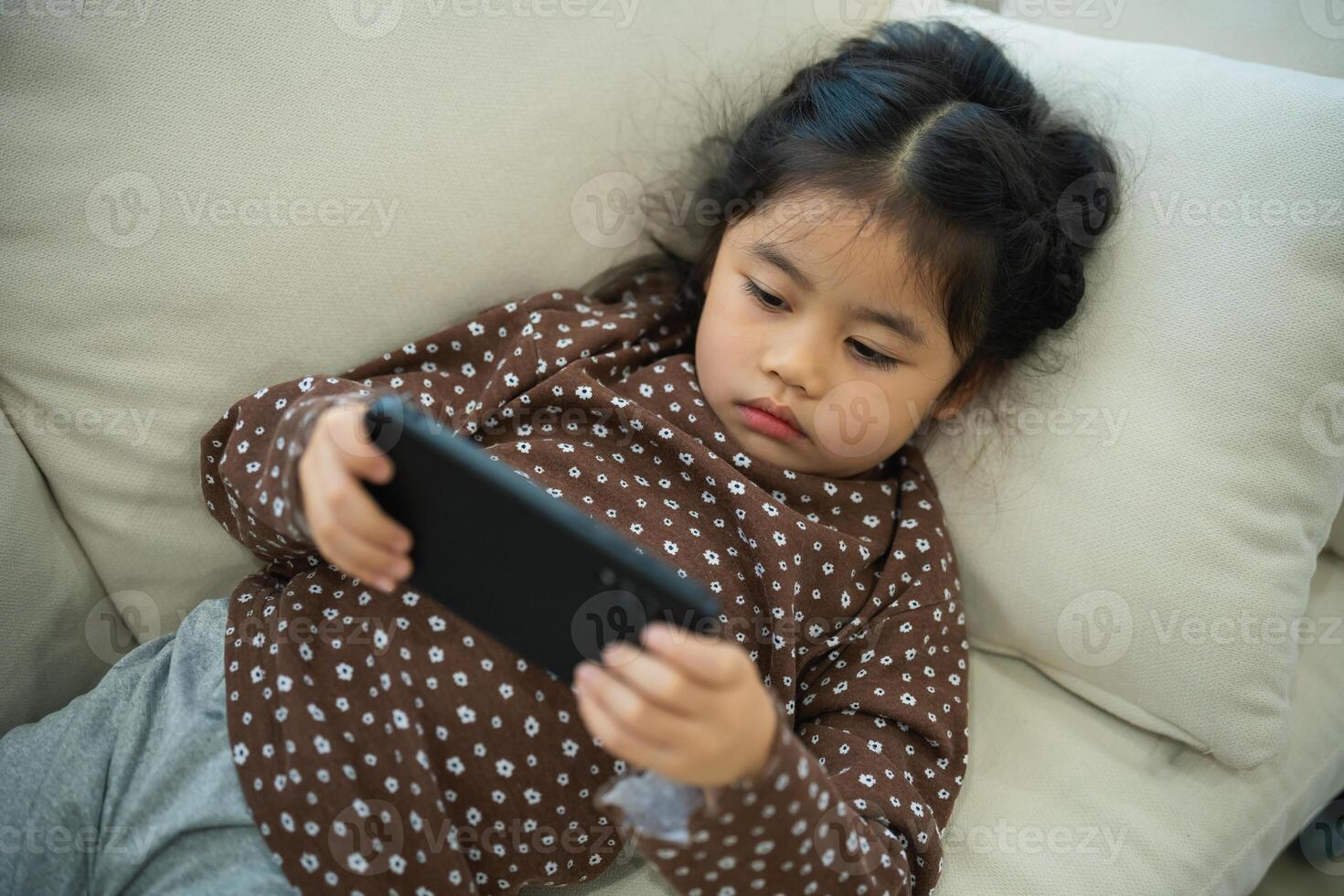 Asian child girl serious or concentrate and using mobile phone screen on couch sofa. Baby smiling funny time use mobile phone. Too much screen time. Girl watching videos while tv, Internet addiction. photo