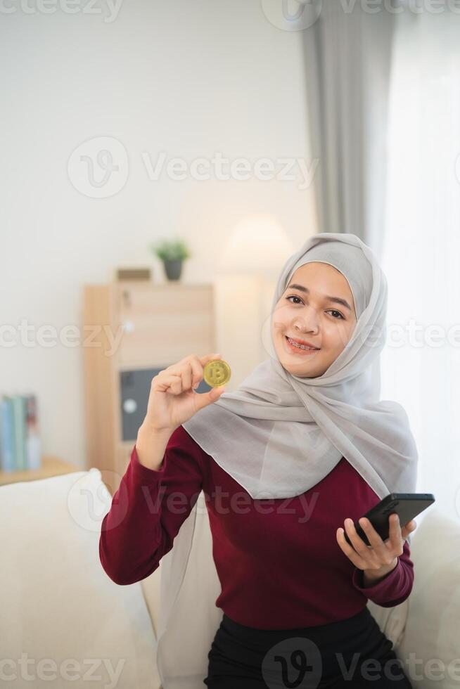 Muslim islam Asian woman wearing hijab smiling holding gold bitcoin crypto currency blockchain sitting on sofa in living room at home. Crypto currency blockchain Saving investment wealth concept. photo