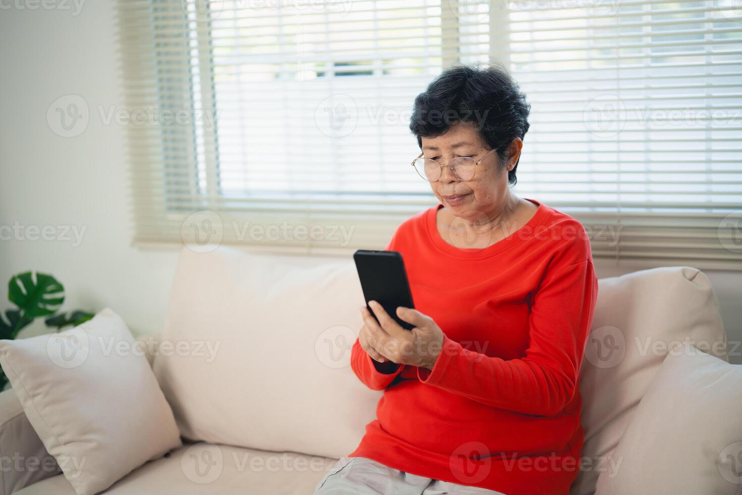 Happy senior old asian woman enjoying using mobile apps texting typing messages sit on sofa, smiling old lady holding smartphone looking smartphone browsing social media or learning technology at home photo