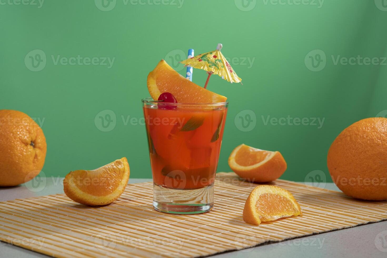 Red cocktail in glass glass filled with ice and mint leaves garnished with orange slice, cherry, sorbet and yellow umbrella on a bamboo napkin with orange pieces photo