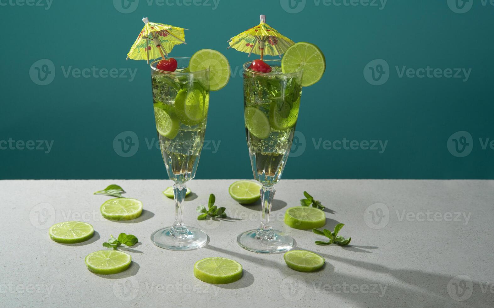 Two cocktails in long glass filled with ice and lemon slices garnished with small yellow umbrellas, cherries and lemon slices on a white table photo