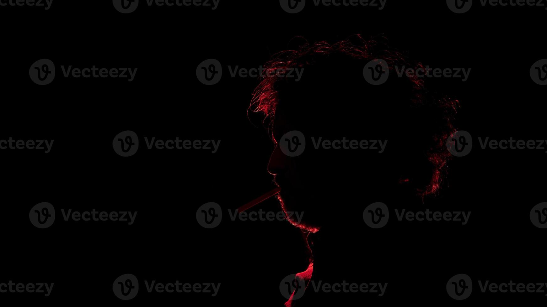 man with a cigarette in his mouth seen in profile in silhouette with red light illuminating his profile against black background photo