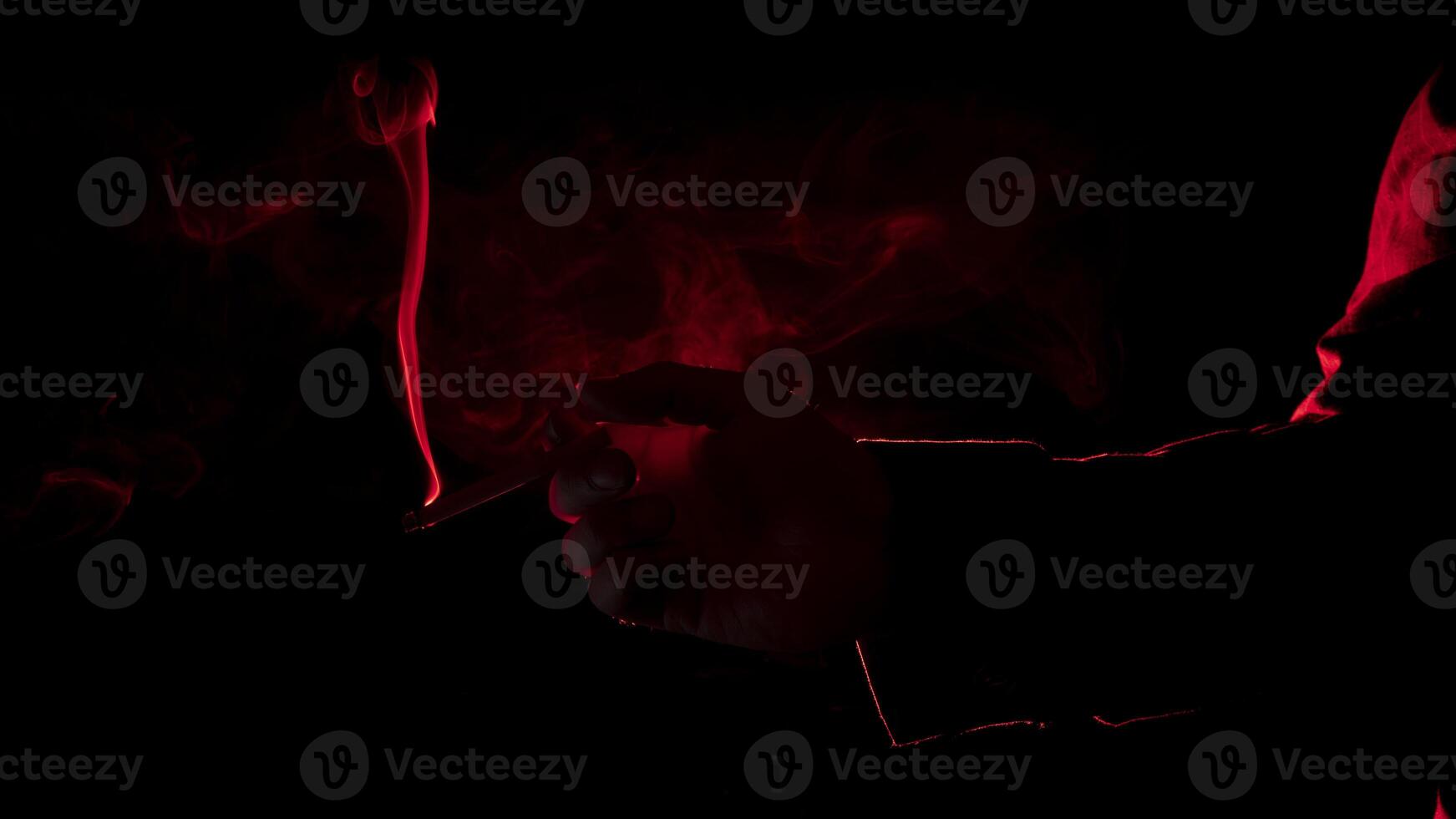 man's hand with a lit cigarette, blowing smoke, seen from profile in silhouette with red light illuminating his profile against black background photo