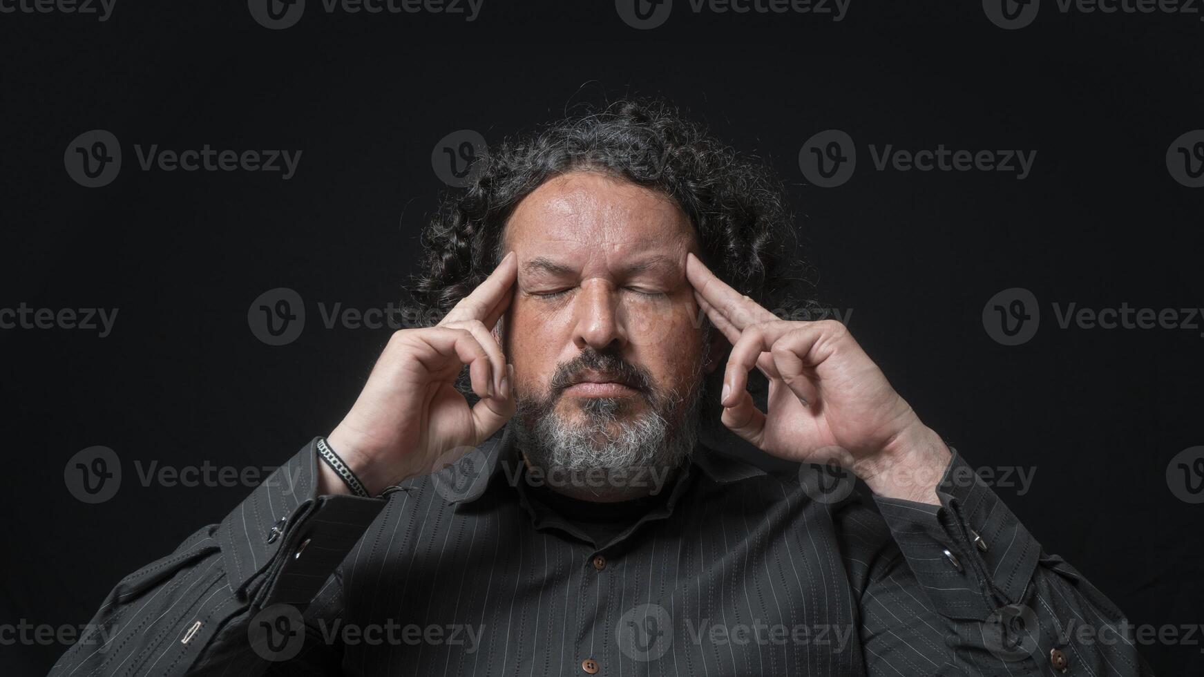 hombre con blanco barba y negro Rizado pelo con concentrado expresión, con su dedos en su templos en su mano, vistiendo negro camisa en contra negro antecedentes foto