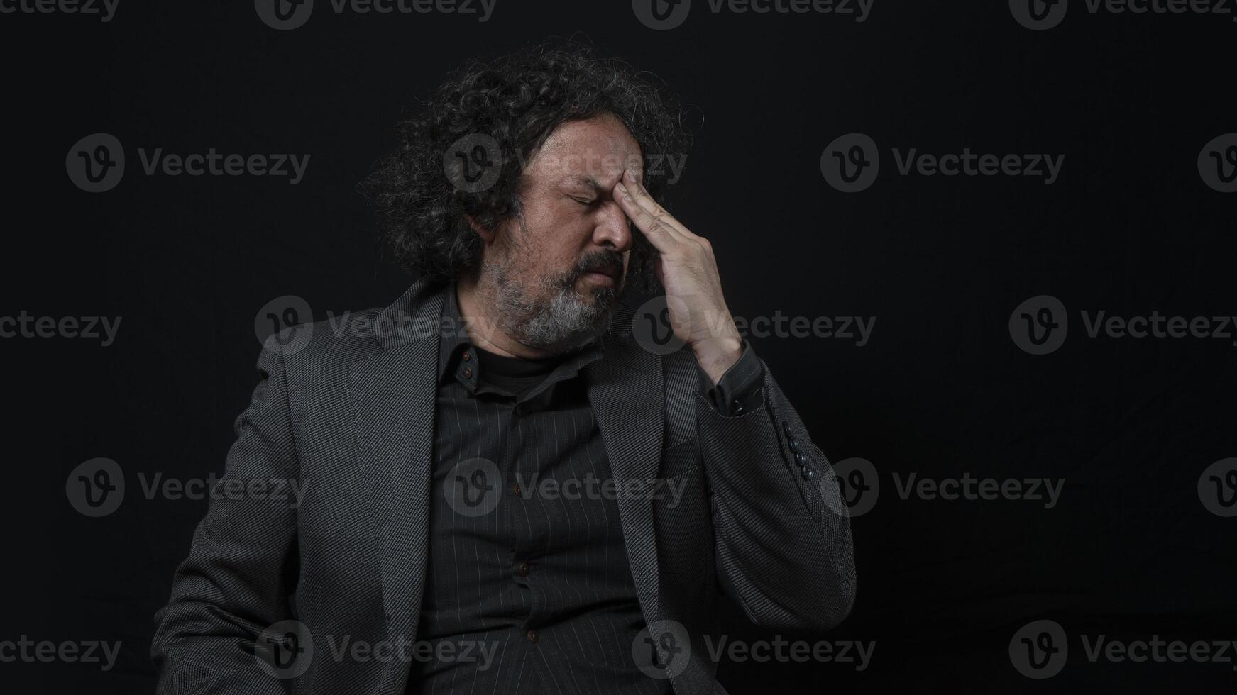 hombre con blanco barba y negro Rizado pelo con dolorido expresión, con su mano en su frente, vistiendo negro camisa en contra negro antecedentes foto