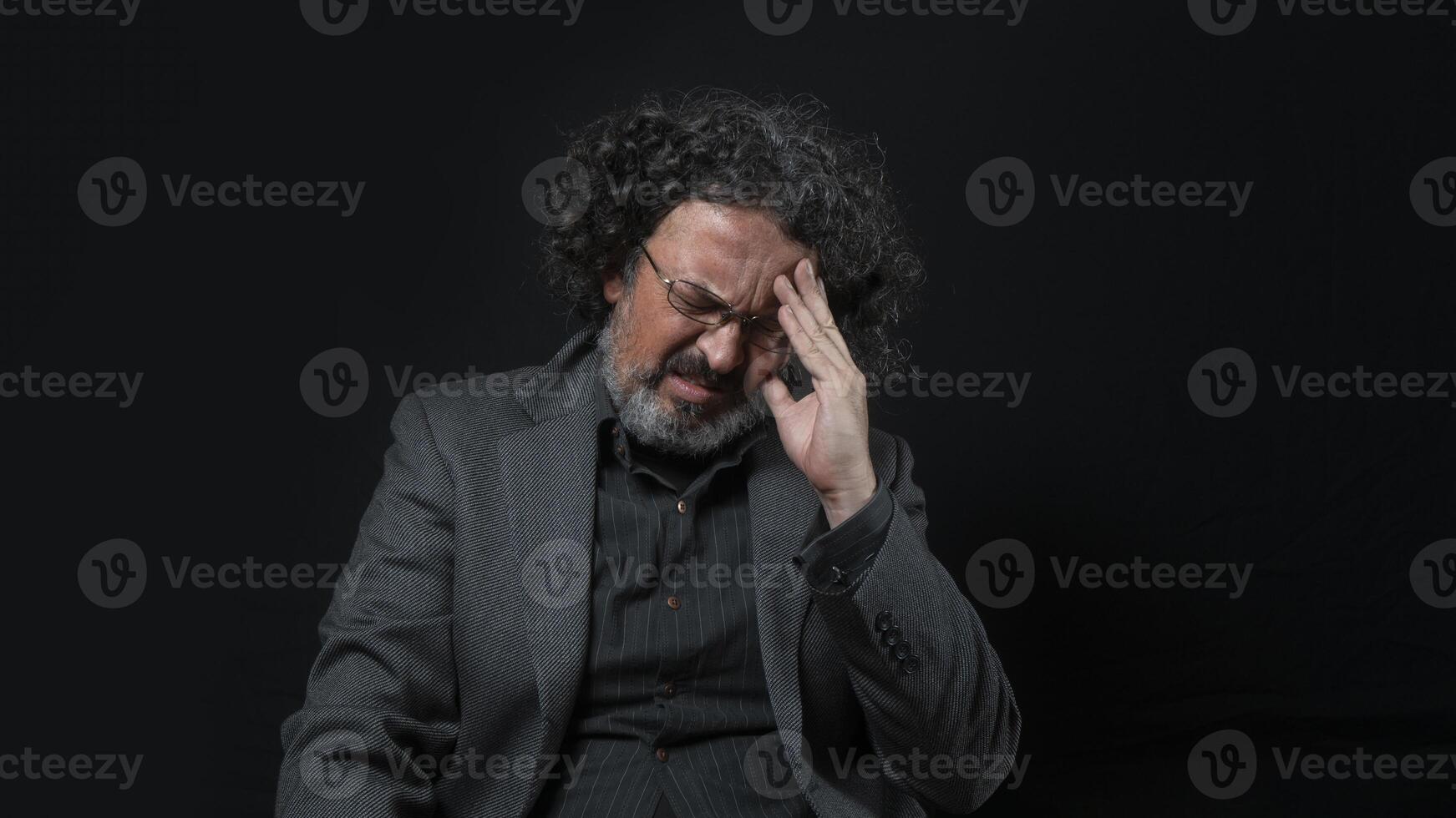 hombre con blanco barba y negro Rizado pelo con dolorido expresión, con su mano en su frente, vistiendo negro camisa en contra negro antecedentes foto