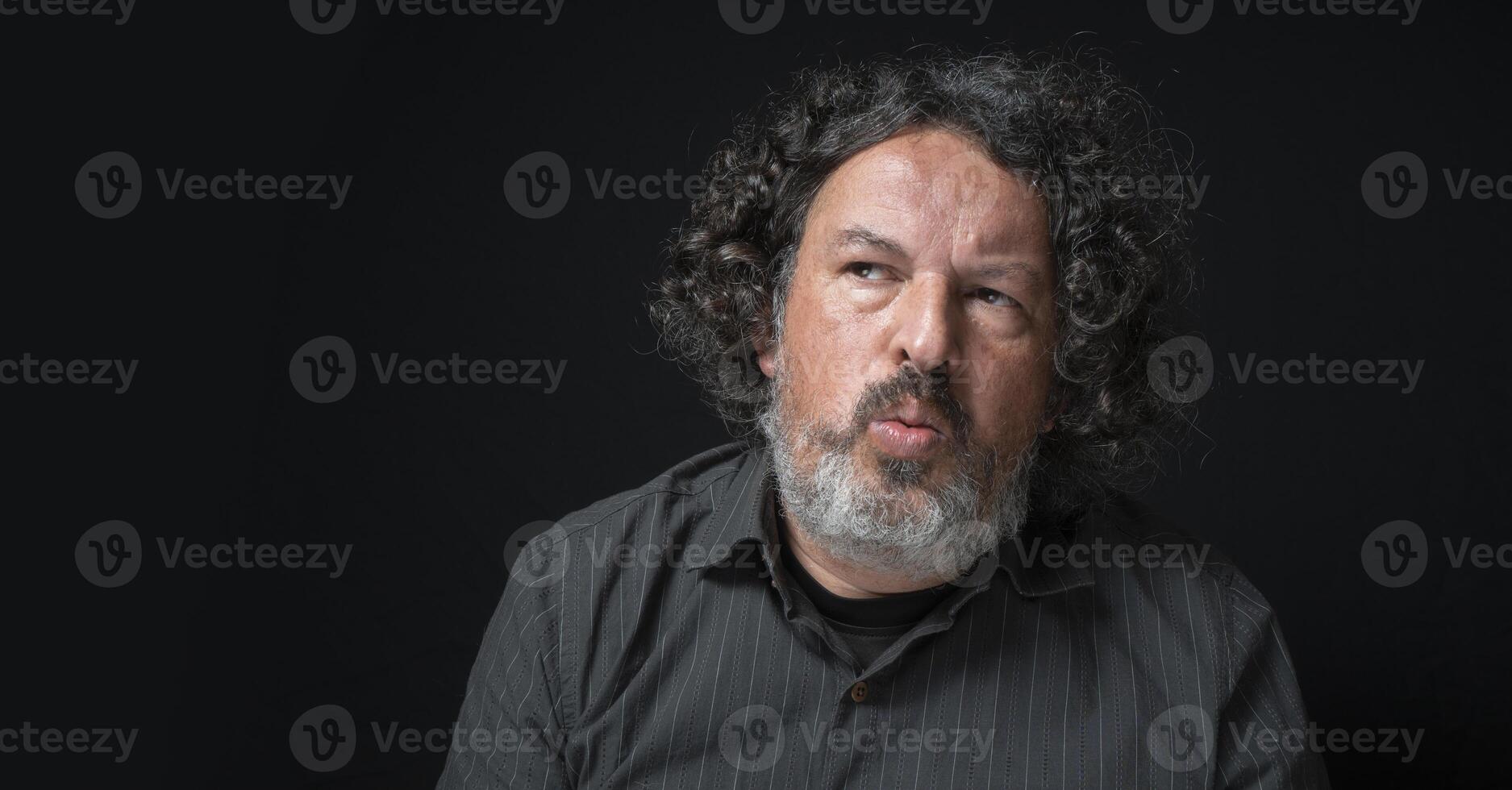 Man with white beard and black curly hair with cautious expression, looking left suspiciously, wearing black shirt against black background photo
