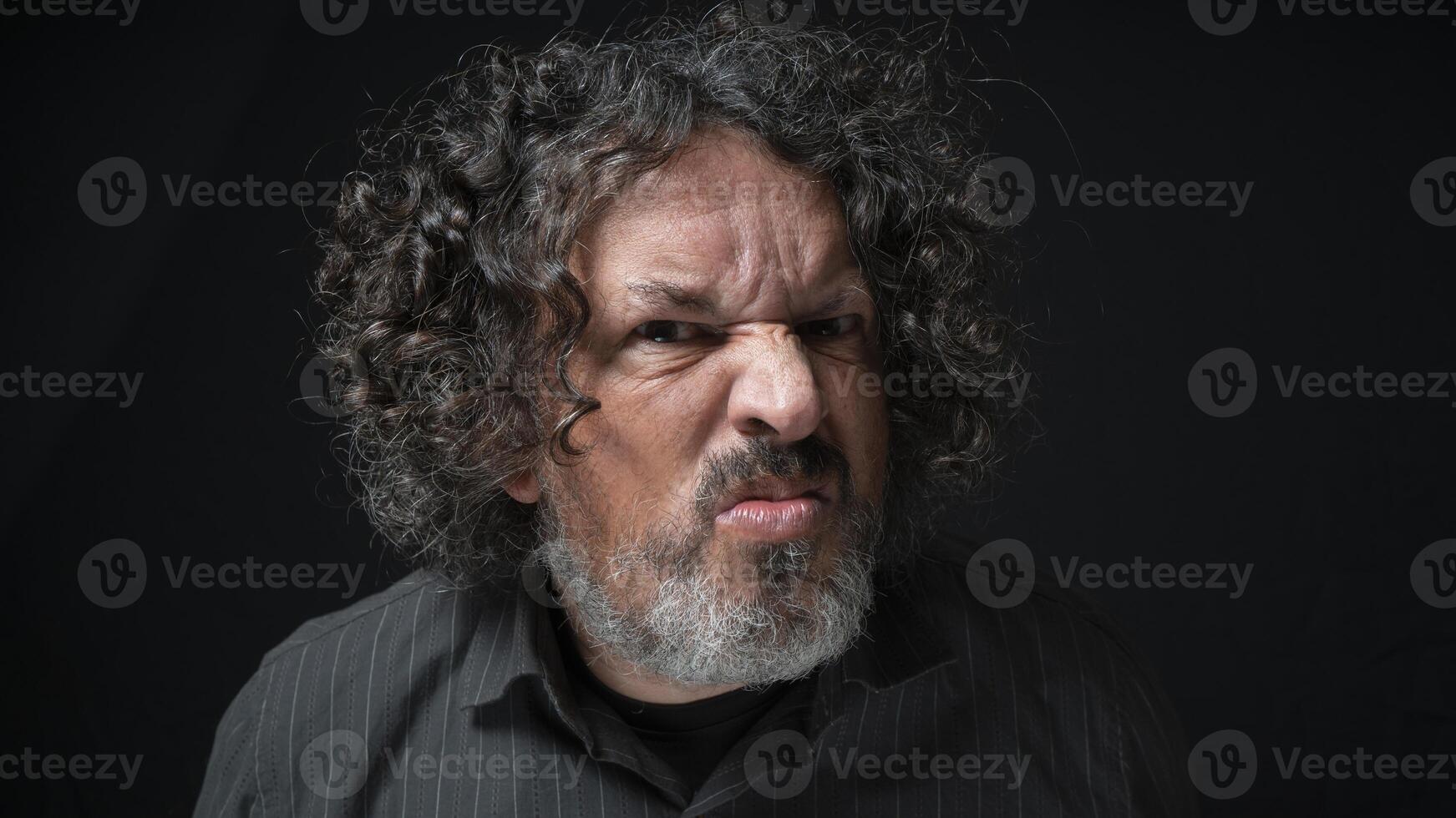 Man with white beard and black curly hair with displeased expression, looking towards camera, wearing black shirt against black background photo