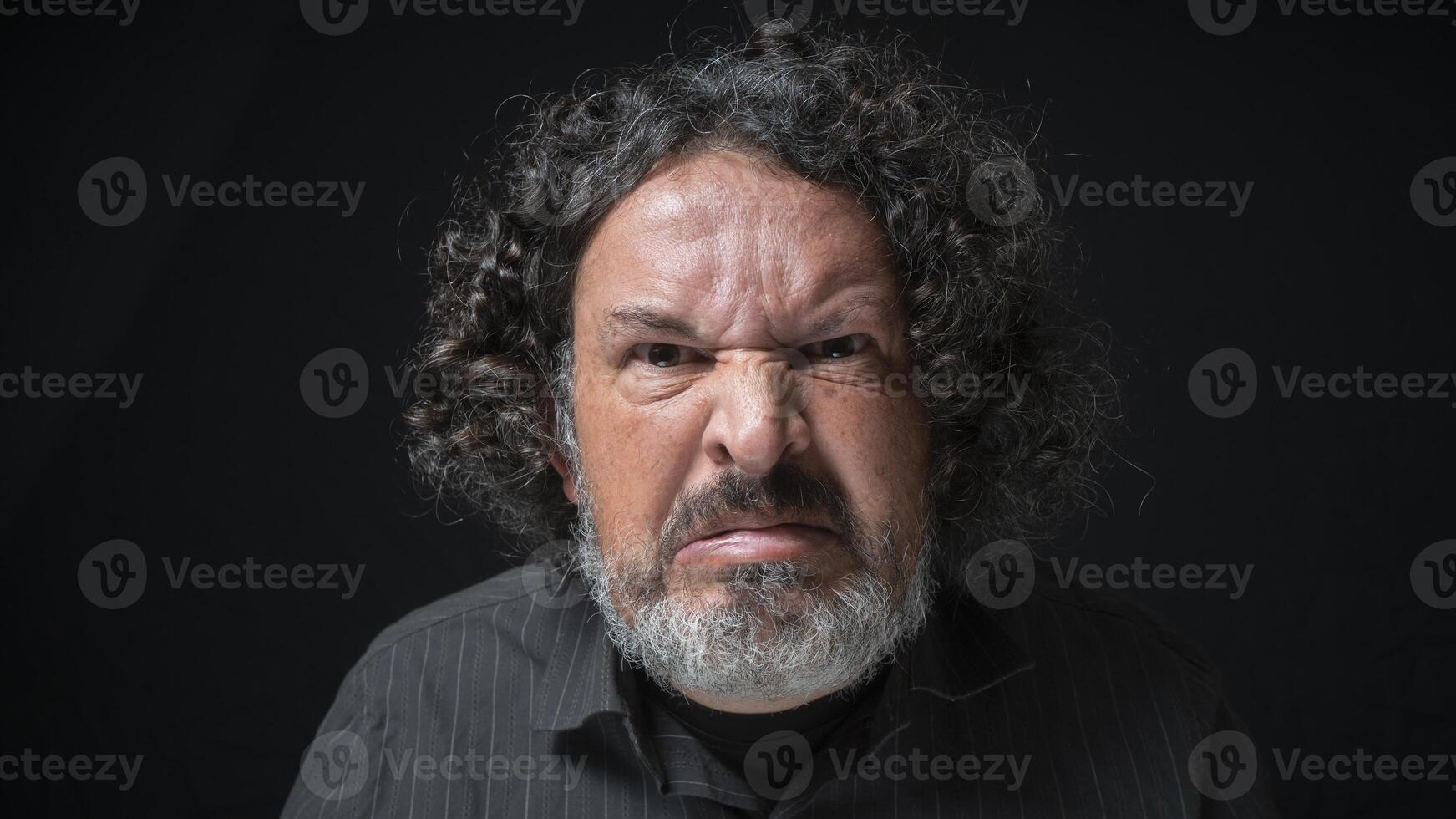 Man with white beard and black curly hair with angry expression, looking straight at camera, wearing black shirt against black background photo