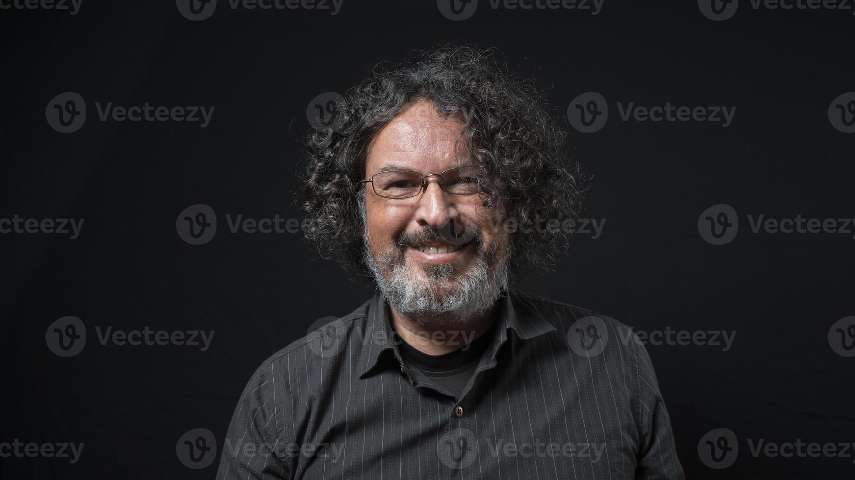 Man with white beard and black curly hair with happy expression, seen from front, wearing black shirt against black background photo