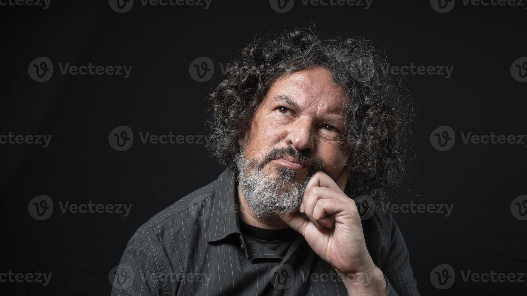 Man with white beard and black curly hair with thoughtful expression, resting his head on his hand, wearing black shirt against black background photo