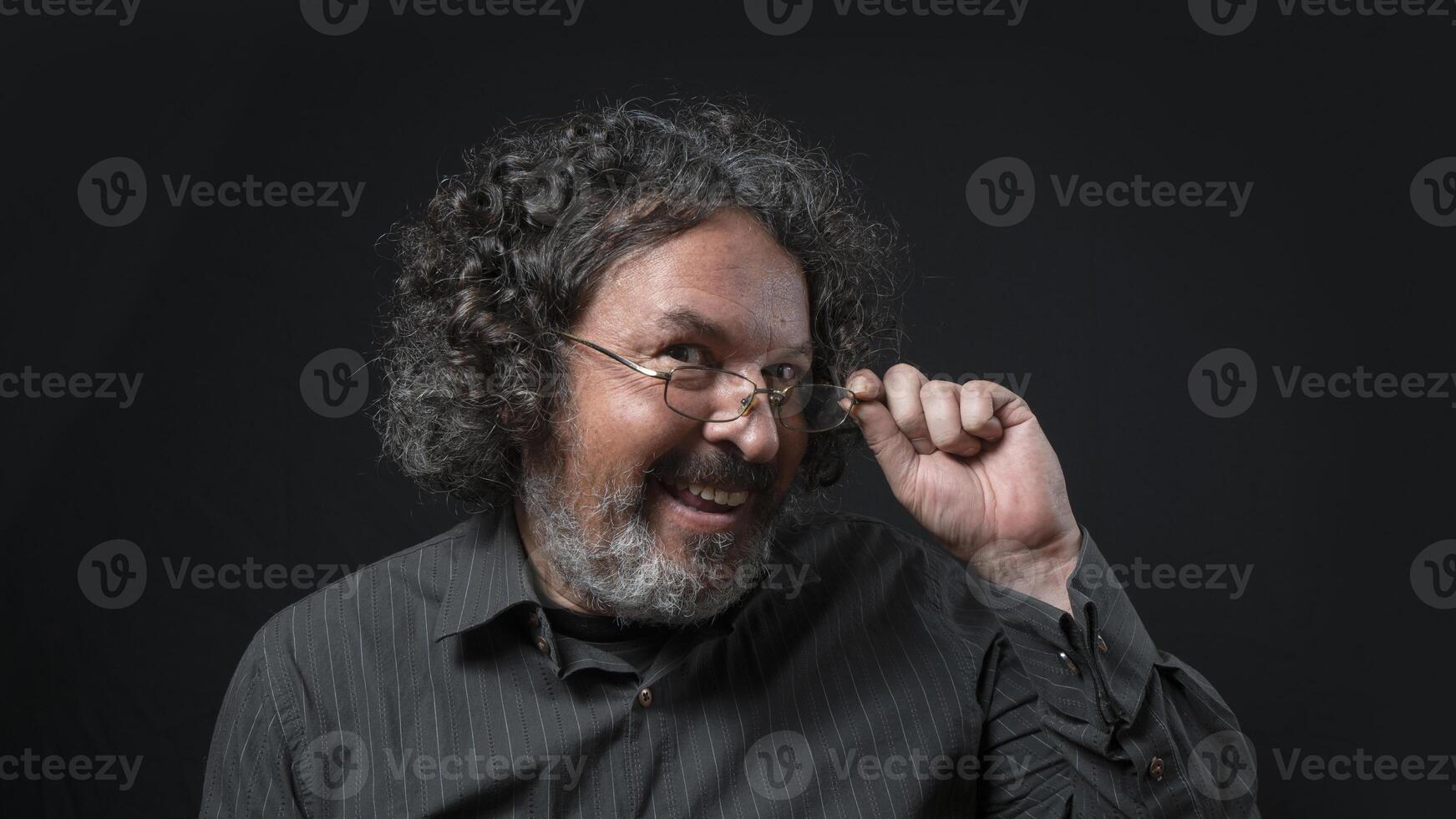 Man with white beard and black curly hair with flirtatious expression, holding his glasses, wearing black shirt against black background photo