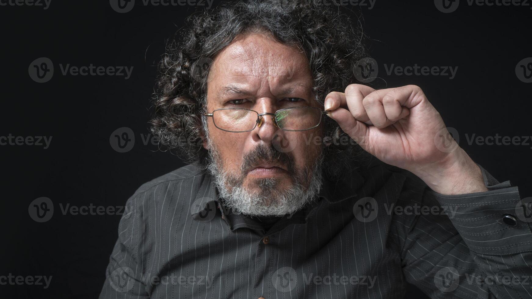 hombre con blanco barba y negro Rizado pelo con curioso expresión, mirando a cámara con recelo, vistiendo negro camisa en contra negro antecedentes foto