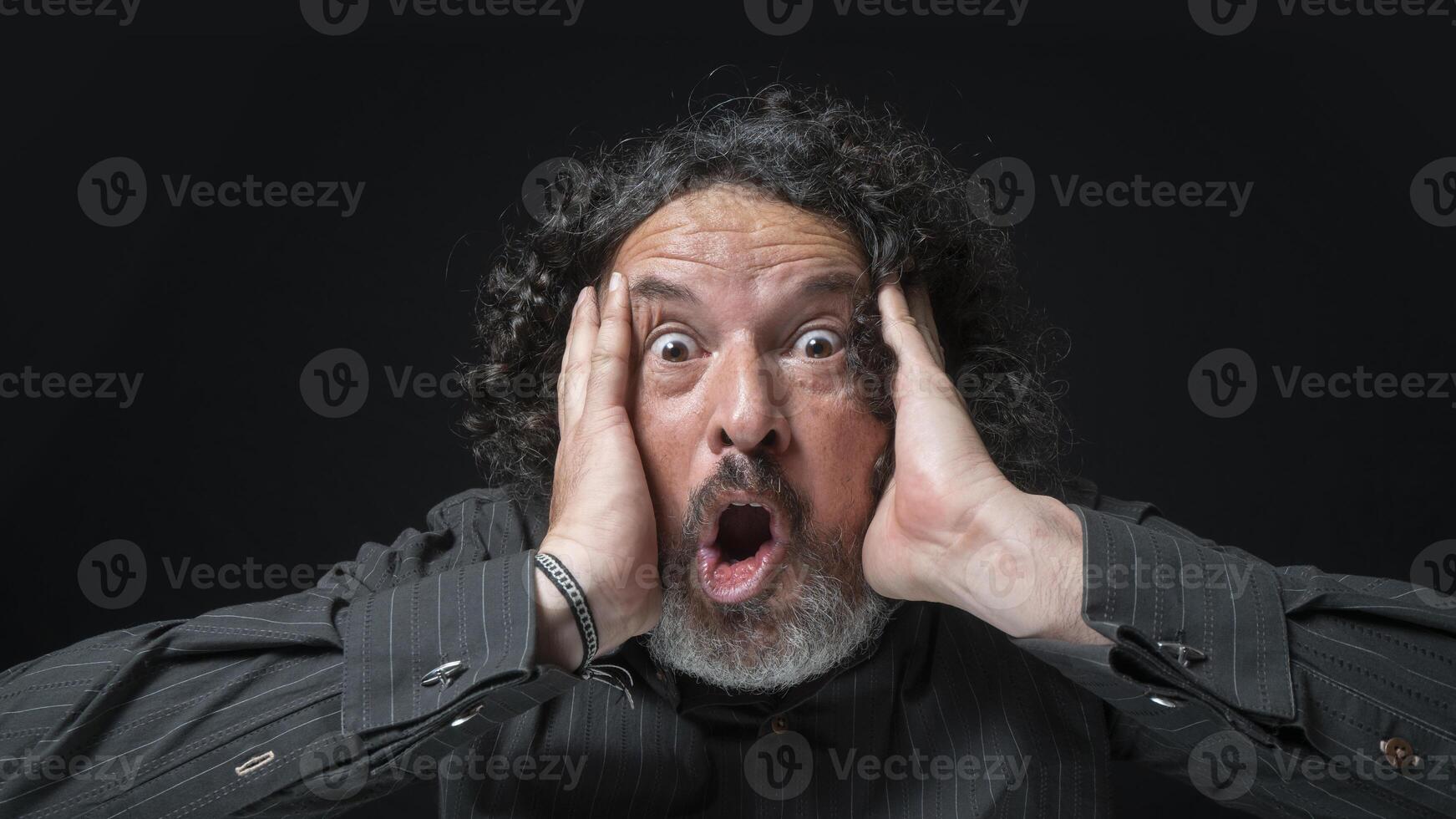 hombre con blanco barba y negro Rizado pelo con sorprendido expresión, manos en rostro, vistiendo negro camisa en contra negro antecedentes foto