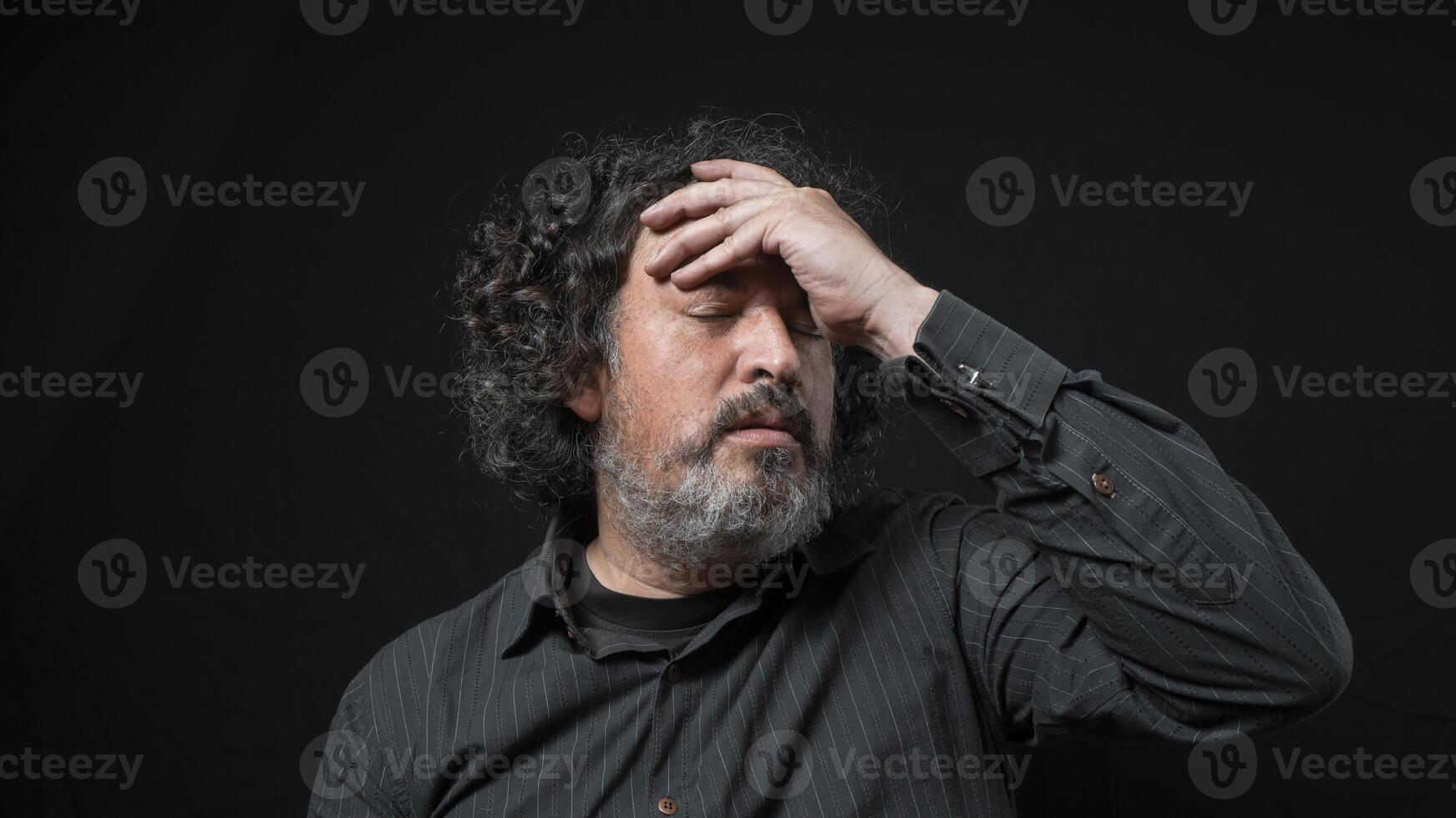 Man with white beard and black curly hair with worried expression, with eyes closed and hand on forehead, wearing black shirt against black background photo