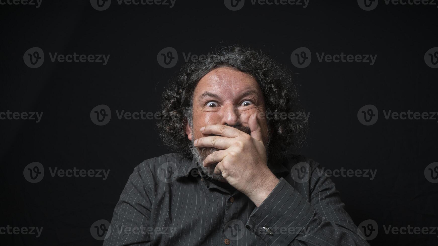 hombre con blanco barba y negro Rizado pelo con gracioso expresión, cubierta su boca con mano, vistiendo negro camisa en contra negro antecedentes foto