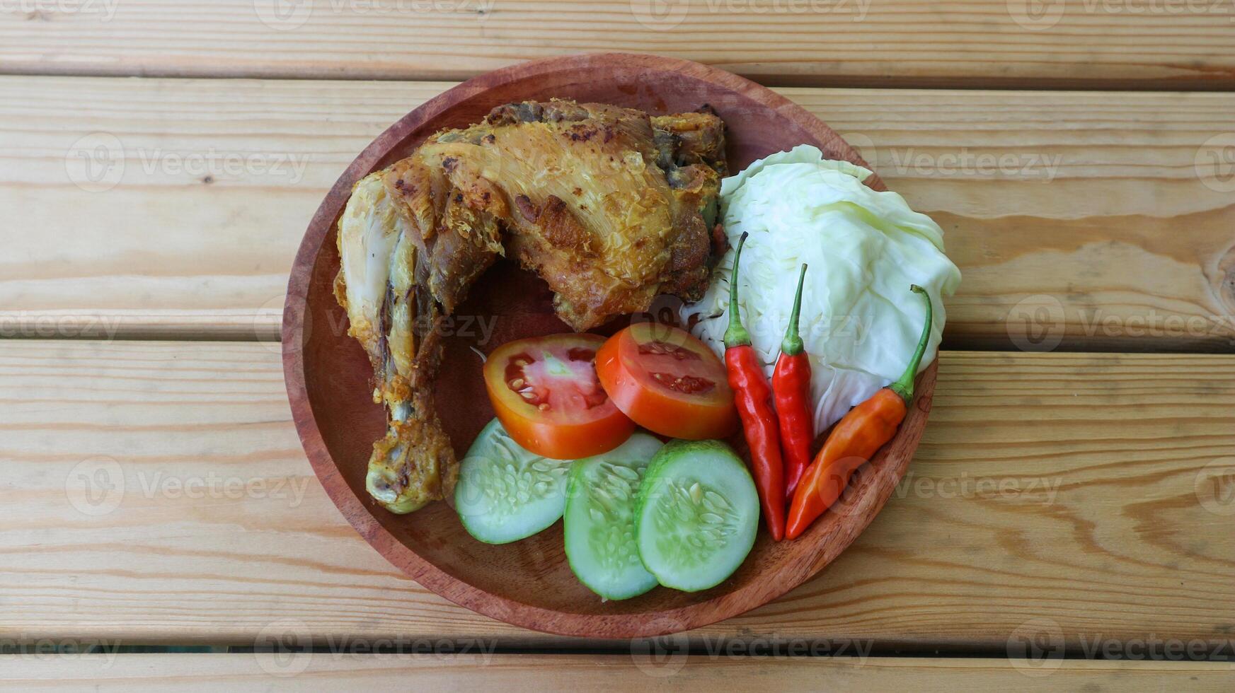 frito pollo con lalapan, frito pollo con Fresco verduras, tomate rebanadas, Pepino y sambal auténtico receta de indonesio pollo lalapán. frito pollo en un de madera mesa. foto