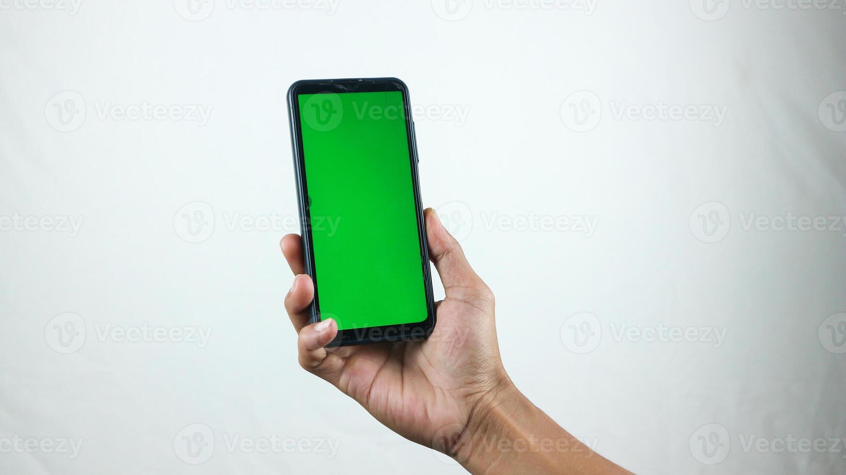 Asian man holding big smartphone with white, red, green, blue blank screen in hand, showing close to camera and pointing at device. Gadget with empty free space for mock up, banner photo