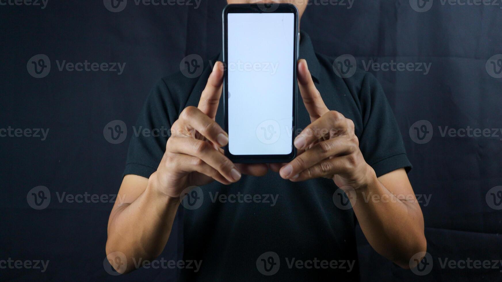 Asian man holding big smartphone with blank screen in hand, showing close to camera and pointing at device. Gadget with empty free space for mock up, banner photo