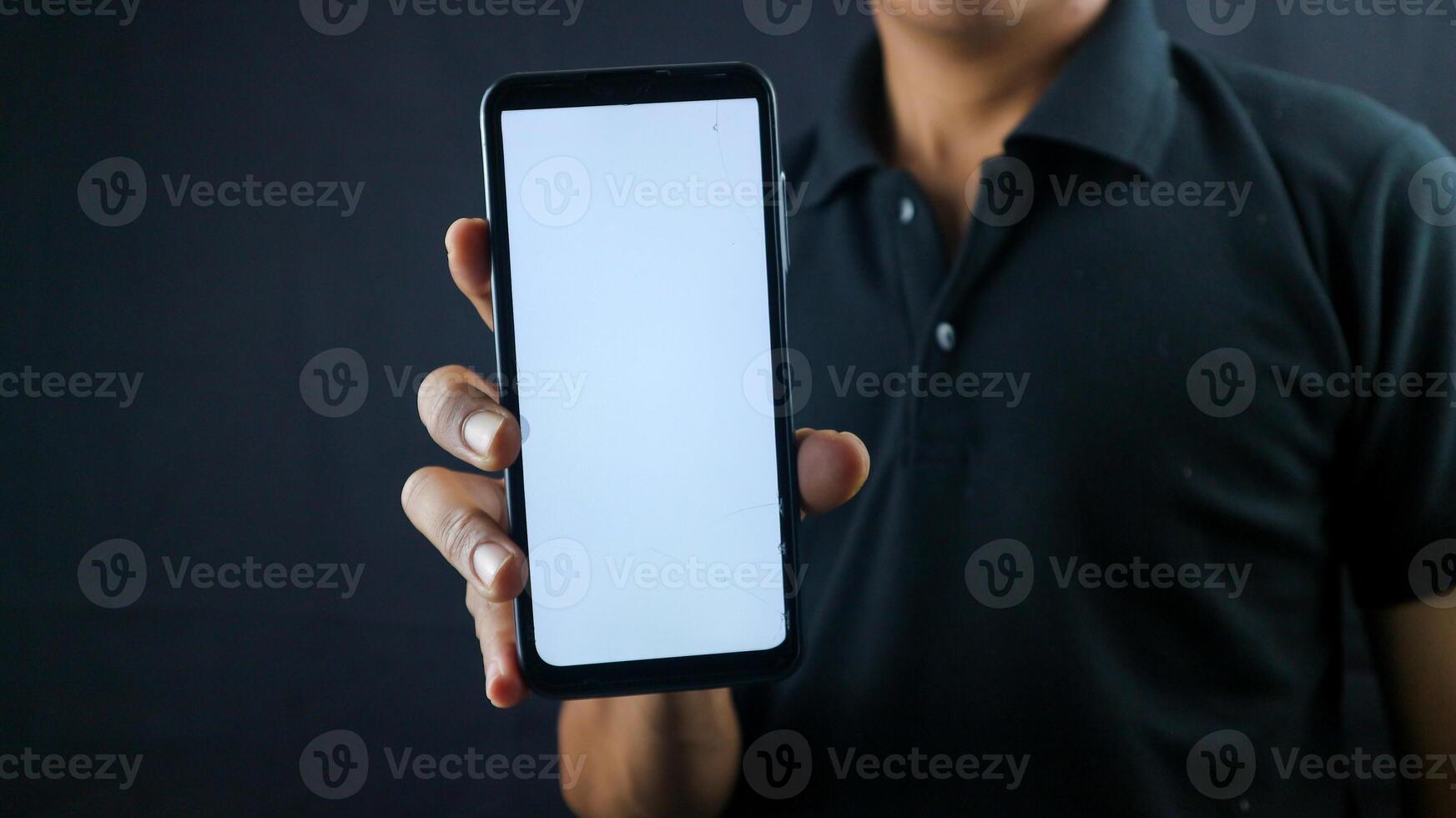 Asian man holding big smartphone with blank screen in hand, showing close to camera and pointing at device. Gadget with empty free space for mock up, banner photo