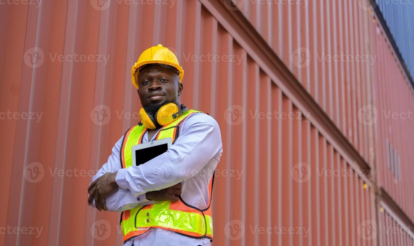 Portrait of African engineer with yellow helmet and safety vest holding digital tablet while standing at cargo container yard photo