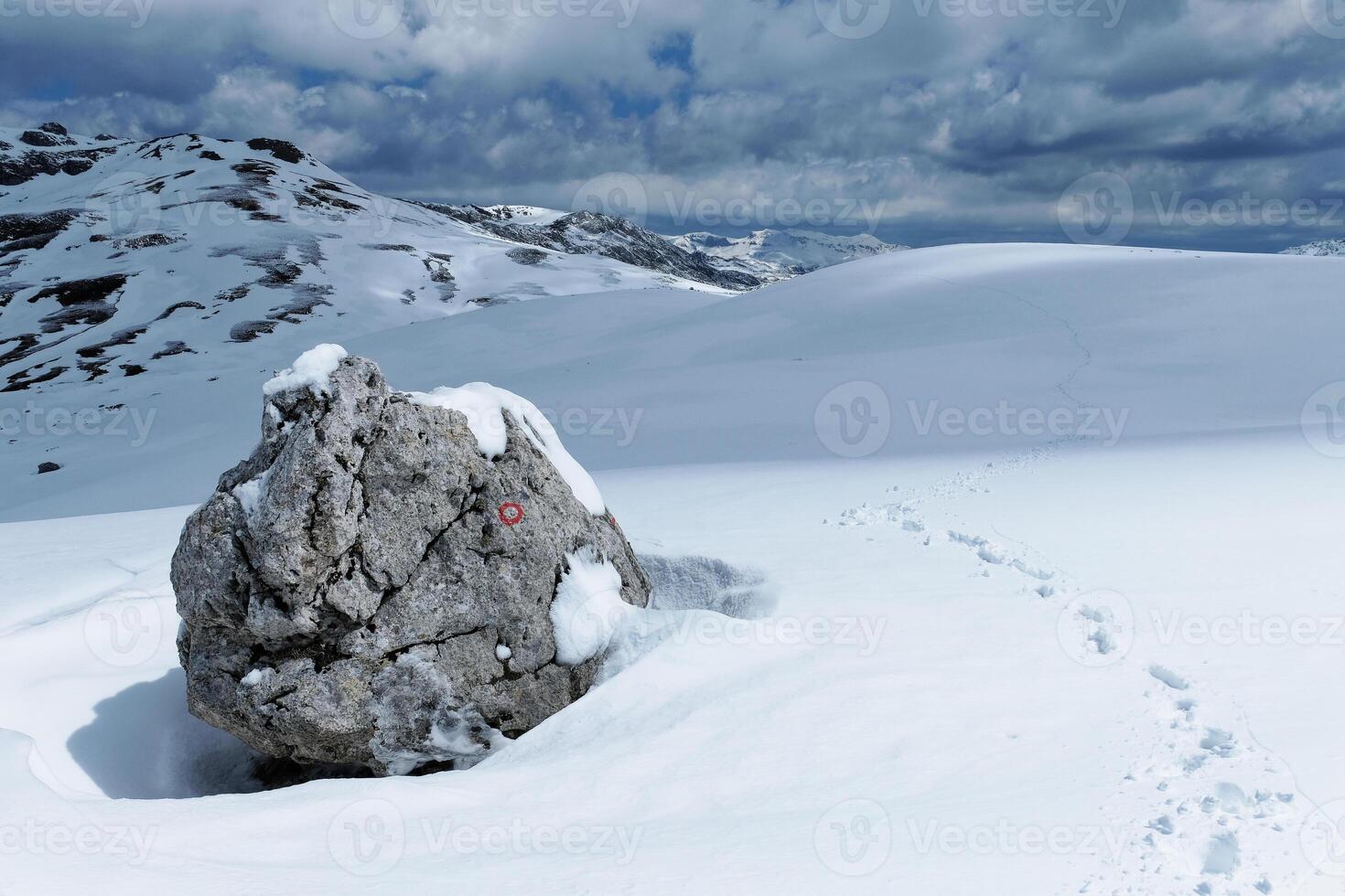 Hiking trail marker in the mountains during winter. Footsteps left by a mountaineer. Snowy white landscape. Red and white mark to follow the path. Hiking and mountaineering concept. photo