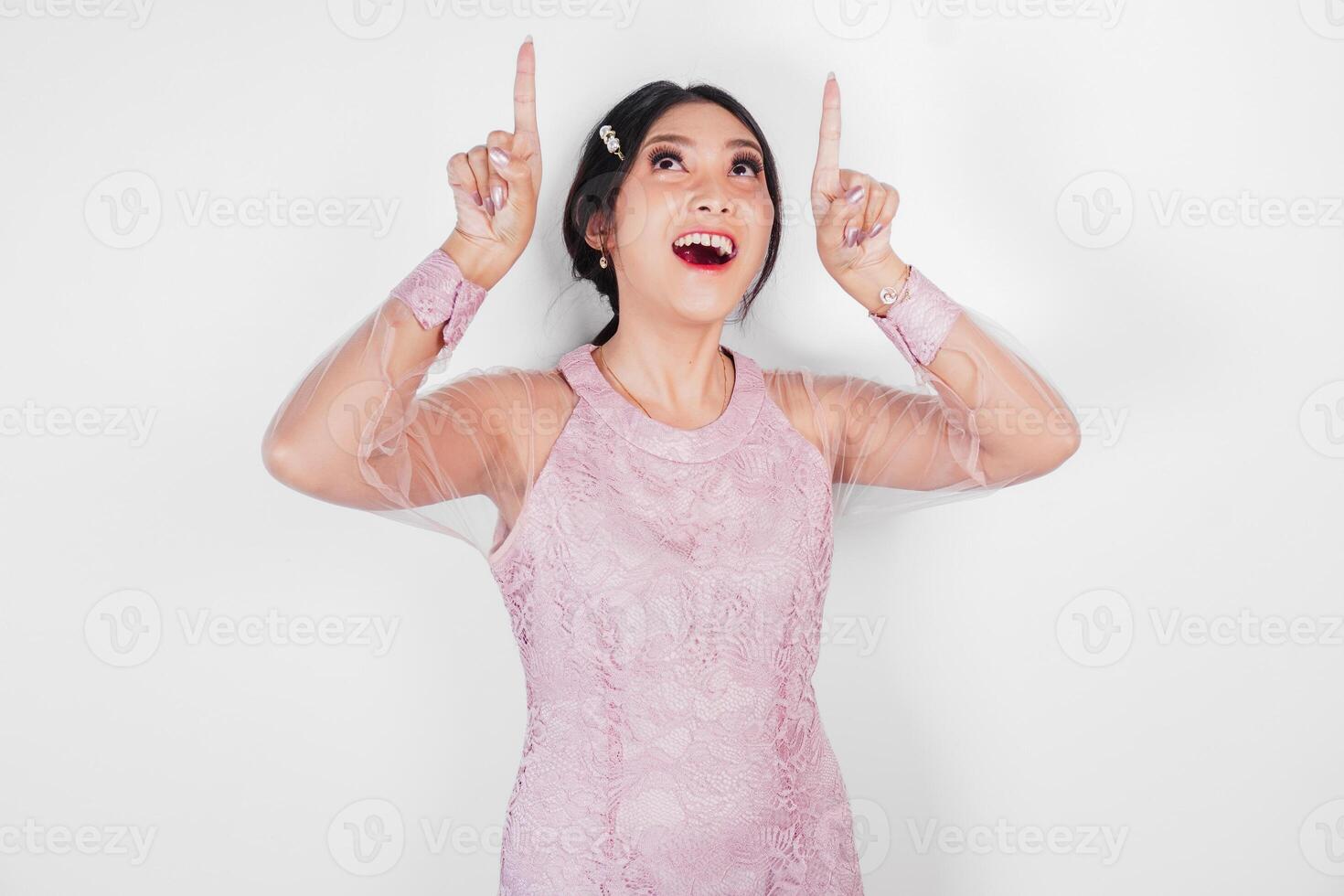 A smiling Asian woman wearing pink dress, pointing to copy space above her, isolated by white background. photo