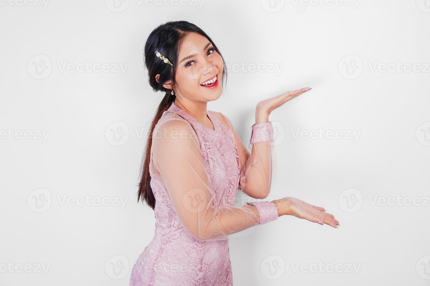 A cheerful Asian woman wearing pink dress is pointing copy space beside her, isolated by white background. photo