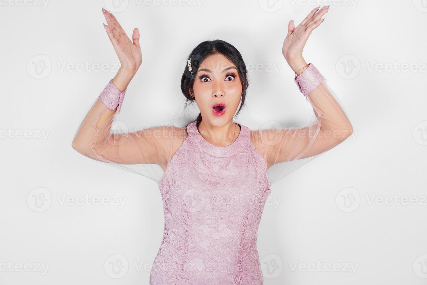 A shocked Asian woman wearing pink dress, pointing to copy space above her, isolated by white background. photo