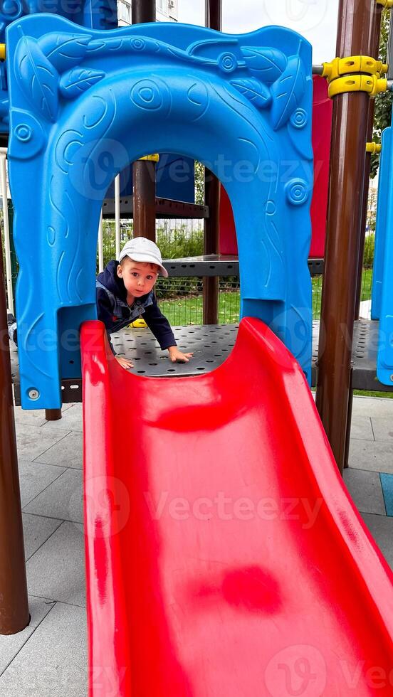 un alegre pequeño chico corredizo en un vistoso patio de recreo equipo. un pequeño chico jugando en un rojo y azul diapositiva foto