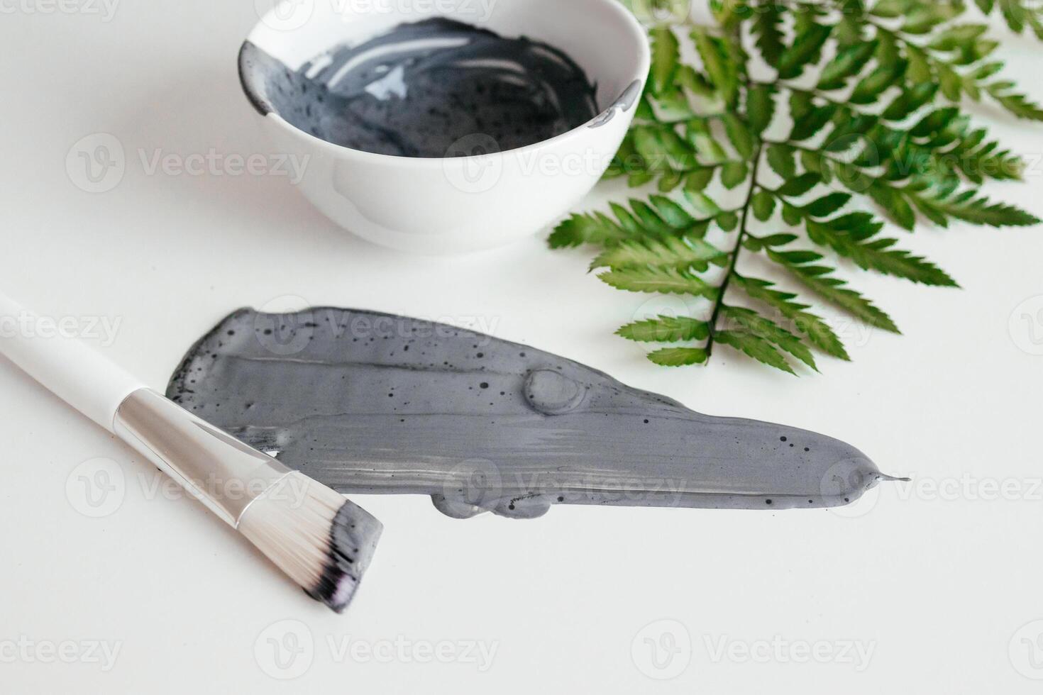 Preparing cosmetic mud mask. Natural wet clay mask with green fern on a white background. Bowl with activated carbon powder, smear texture of clay mud face mask. Spa and skin care concept. photo