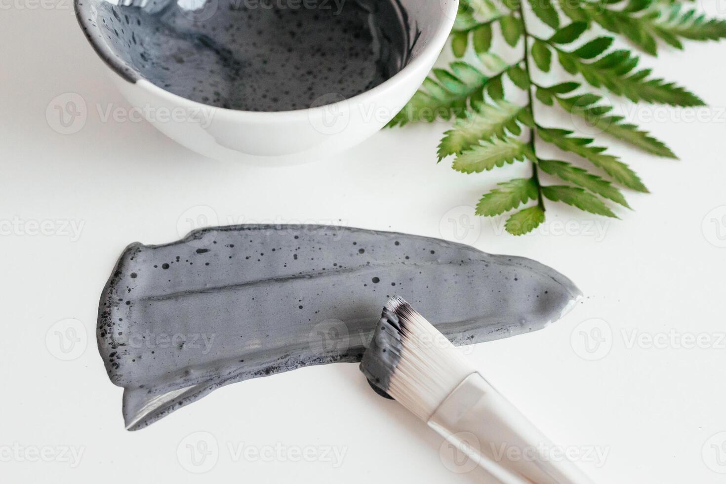 Preparing cosmetic mud mask. Natural wet clay mask with green fern on a white background. Bowl with activated carbon powder, smear texture of clay mud face mask. Spa and skin care concept. photo