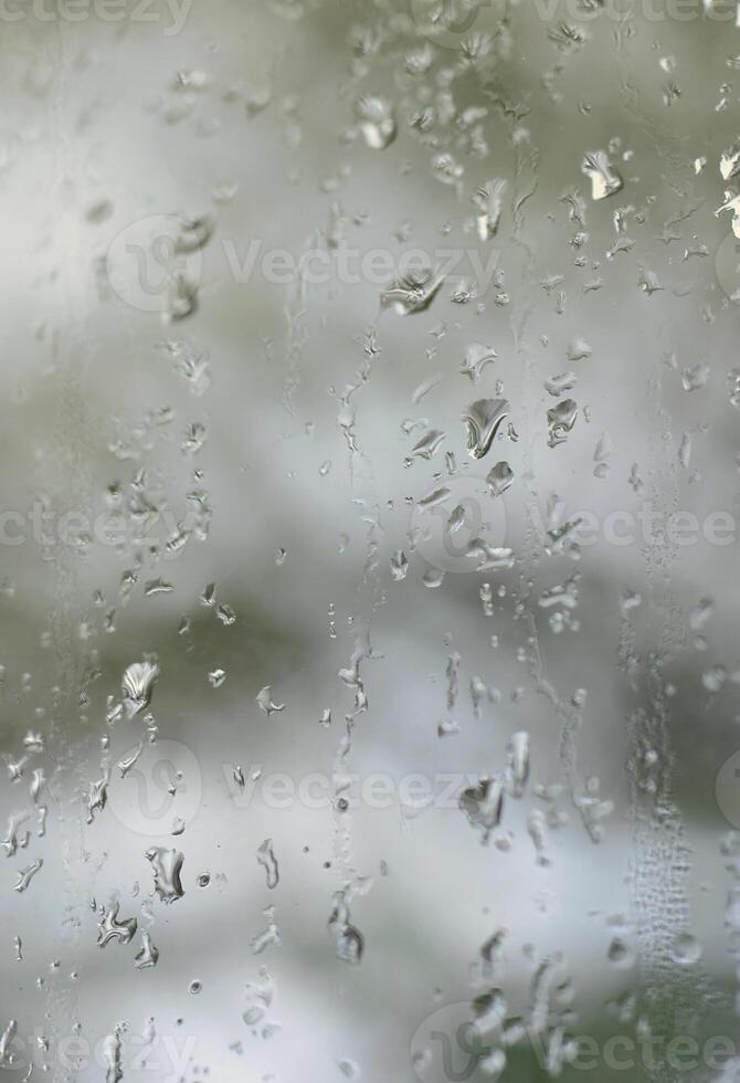 A photo of rain drops on the window glass with a blurred view of the blossoming green trees. Abstract image showing cloudy and rainy weather conditions