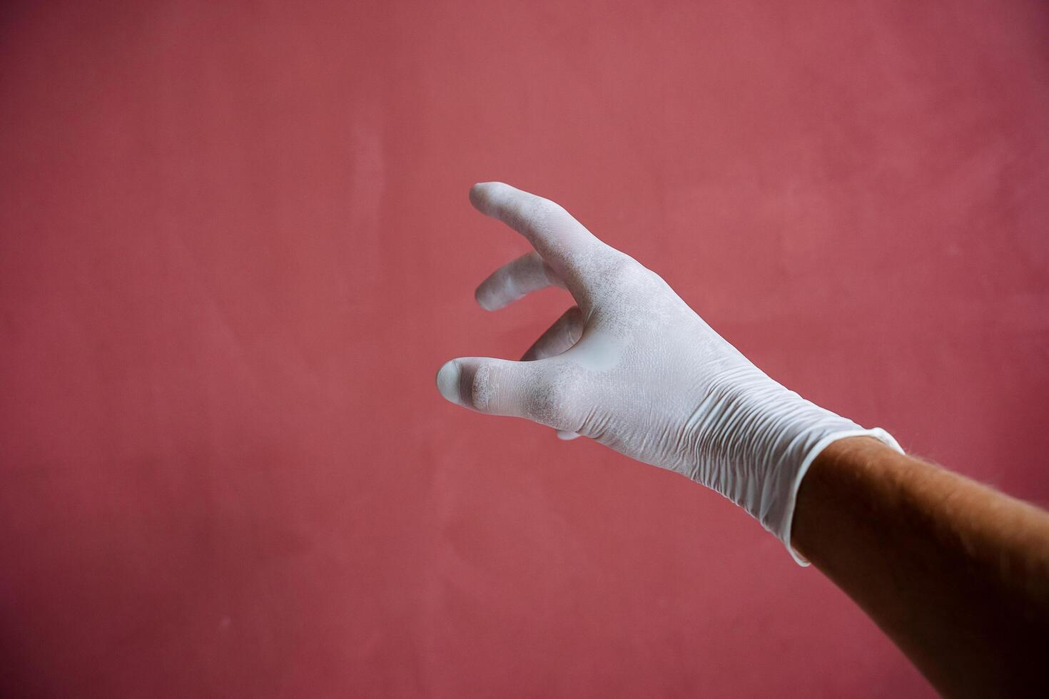 un mano en un blanco médico guante estiramientos adelante, un sudoroso mano debajo látex, dedos untado fuera en contra un granate muro, mano proteccion foto