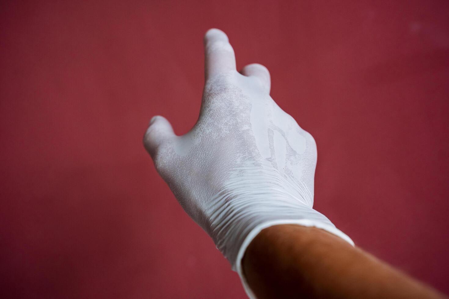 un mano en un blanco médico guante estiramientos adelante, un sudoroso mano debajo látex, dedos untado fuera en contra un granate muro, mano proteccion foto