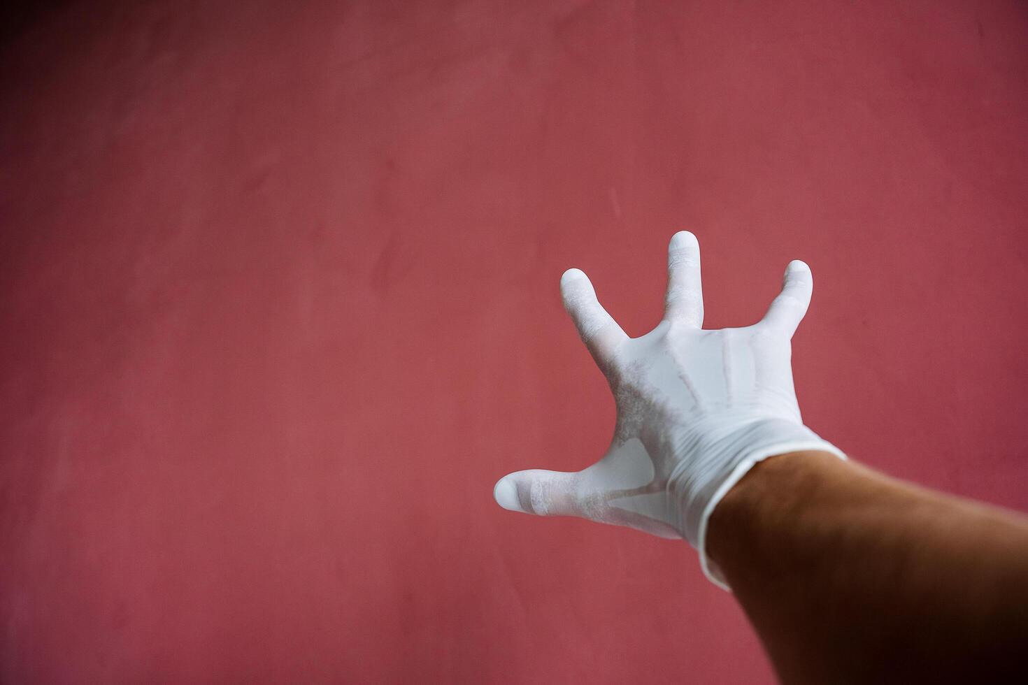 un mano en un blanco médico guante estiramientos adelante, un sudoroso mano debajo látex, dedos untado fuera en contra un granate muro, mano proteccion foto