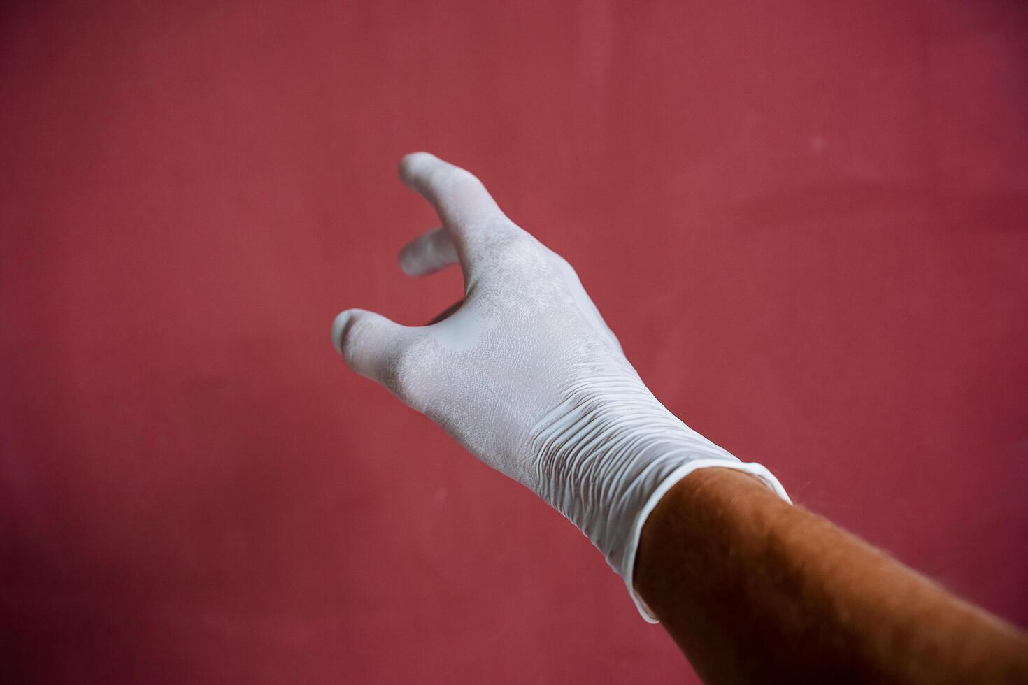 un mano en un blanco médico guante estiramientos adelante, un sudoroso mano debajo látex, dedos untado fuera en contra un granate muro, mano proteccion foto