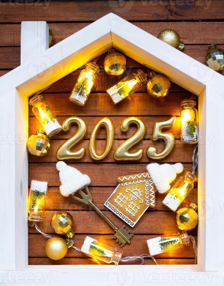 House key with keychain cottage on festive brown wooden background with stars, lights of garlands. New Year 2025 golden letters under the roof. Purchase, construction, relocation, mortgage, insurance photo