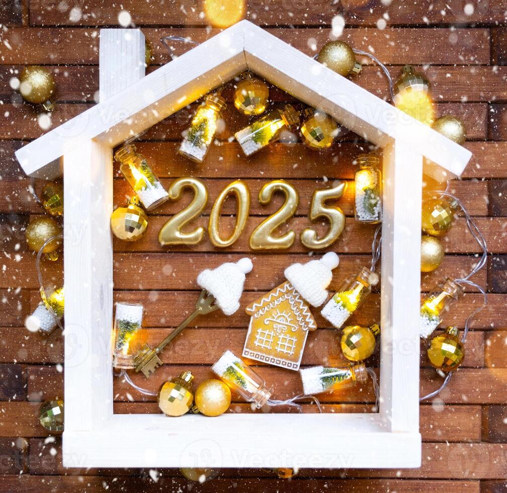 House key with keychain cottage on festive brown wooden background with stars, lights of garlands. New Year 2025 golden letters under the roof. Purchase, construction, relocation, mortgage, insurance photo