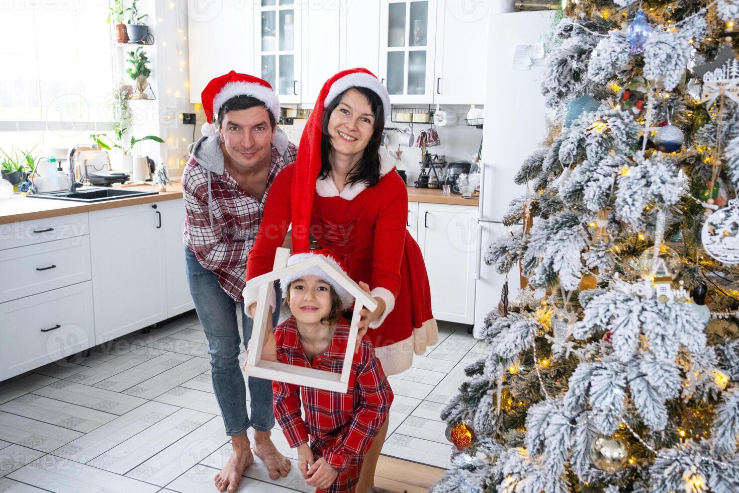 contento familia en Papa Noel sombreros con minúsculo casa en acogedor hogar con Navidad decoración de festivo blanco cocina. regalo para nuevo año. seguro, Moviente a nuevo casa, hipoteca, alquilar y compra real inmuebles foto