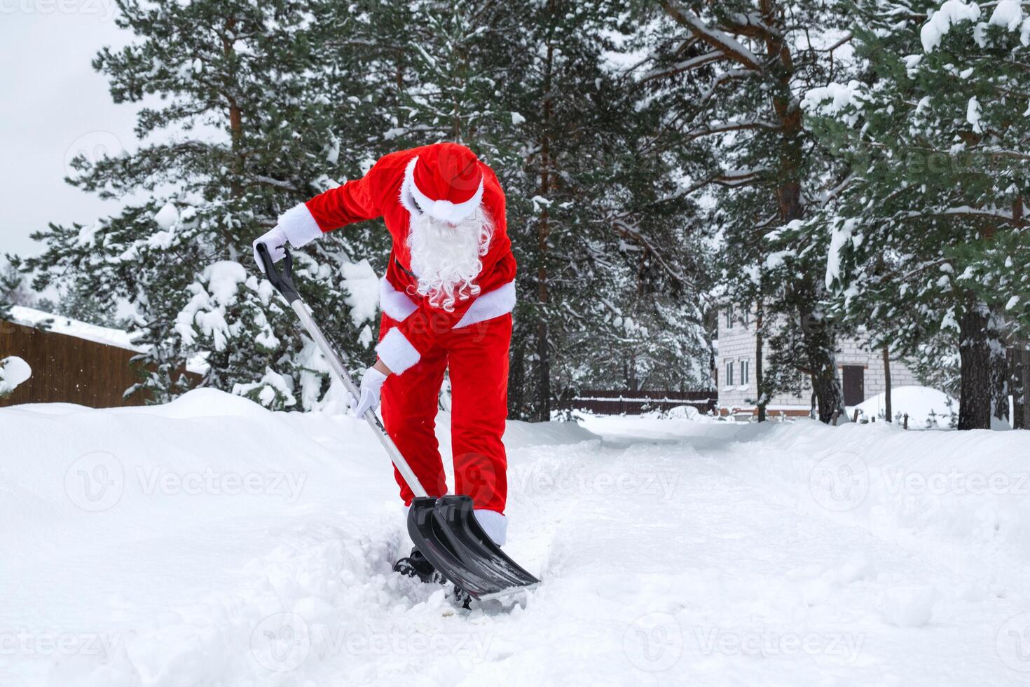 Santa Claus cleans snow with shovel in winter outdoors after a snowfall. Cleaning the streets in the village, clearing the passage for cars, difficult weather conditions for Christmas and New Year photo