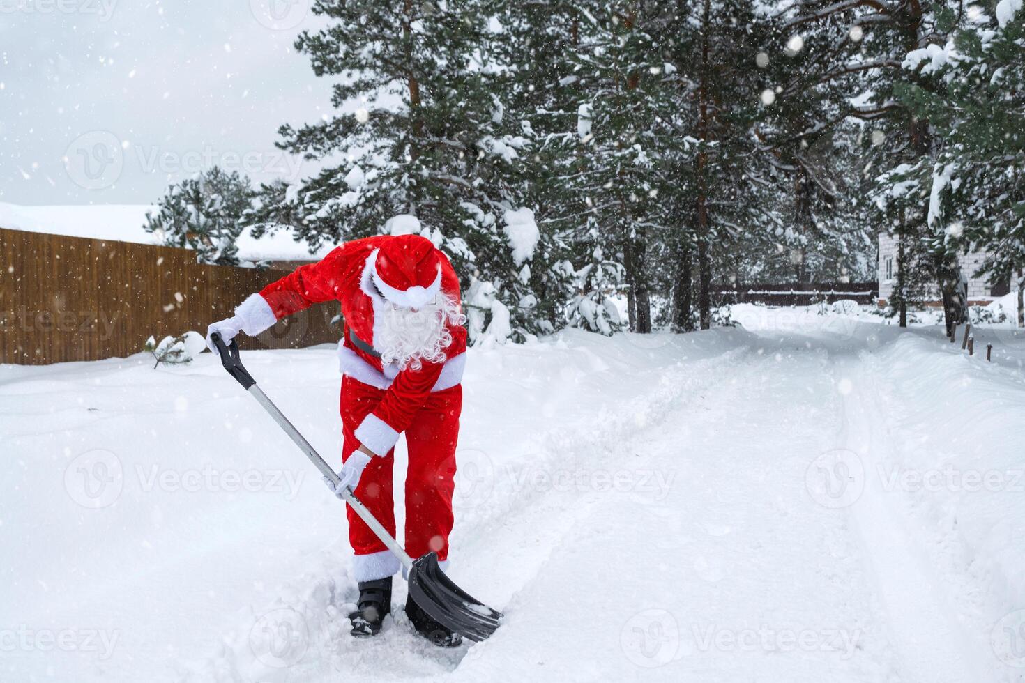 Santa Claus cleans snow with shovel in winter outdoors after a snowfall. Cleaning the streets in the village, clearing the passage for cars, difficult weather conditions for Christmas and New Year photo