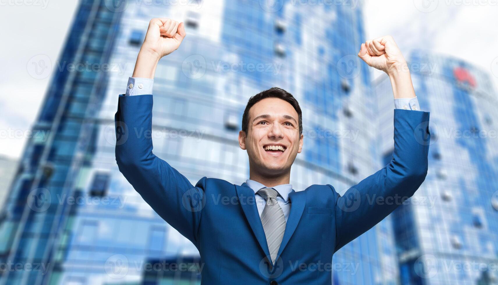 Businessman smiling and raising his fist in the air, with office building in background - business success, achievement, and win concepts photo