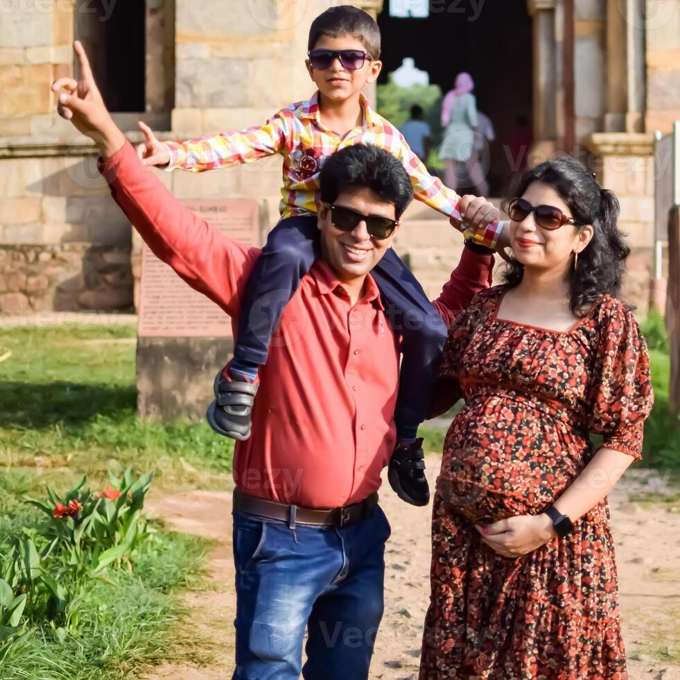 Indian couple posing for maternity baby shoot with their 5 year old kid. The couple is posing in a lawn with green grass and the woman is flaunting her baby bump in Lodhi Garden in New Delhi, India photo