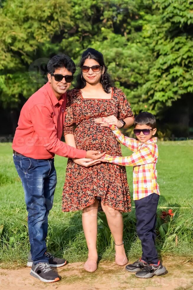Indian couple posing for maternity baby shoot with their 5 year old kid. The couple is posing in a lawn with green grass and the woman is flaunting her baby bump in Lodhi Garden in New Delhi, India photo