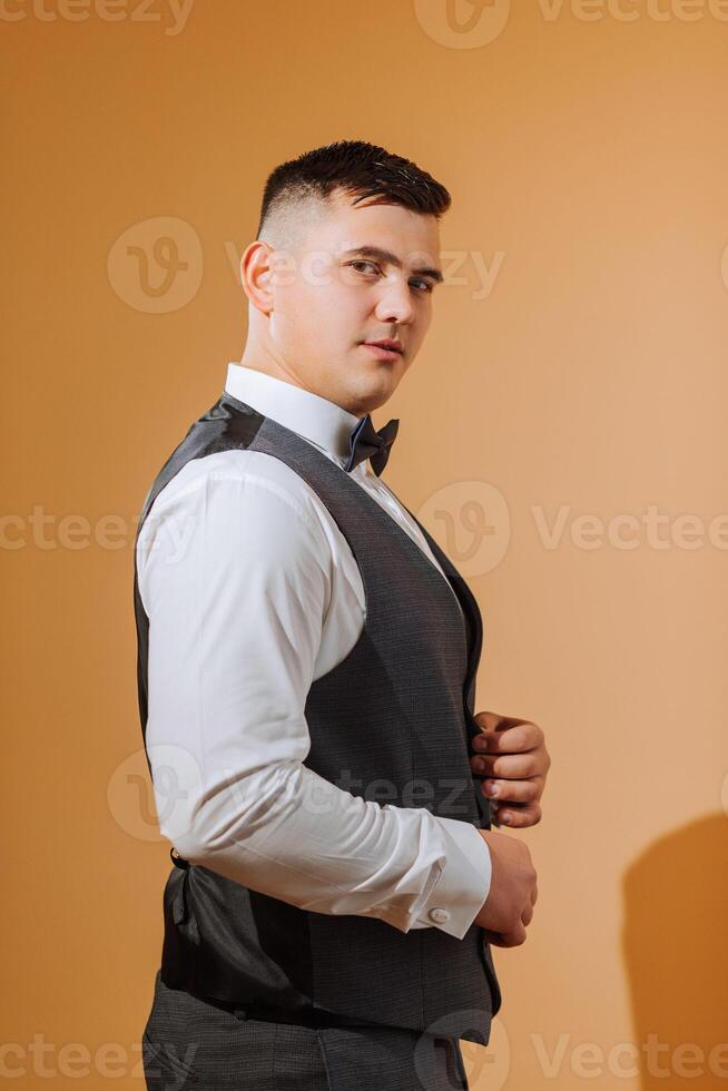 a young smiling man in a white shirt wears a bow tie and a black waistcoat. A groom with a short hairstyle on an orange background puts on his black suit and prepares for the wedding photo