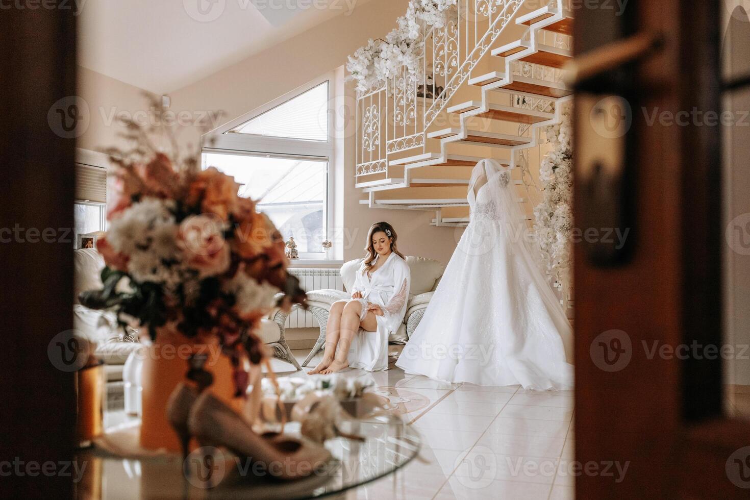 Morning of the bride before the wedding. A beautiful young woman with a veil and a beautiful hairstyle in a white robe near her wedding dress on a mannequin. Professional makeup. Last preparations. photo