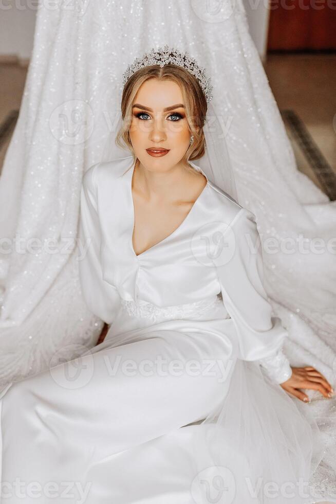 un novia en un seda traje con un velo poses en su habitación, sentado en el piso siguiente a su Boda vestido. Mañana de el novia. preparación foto