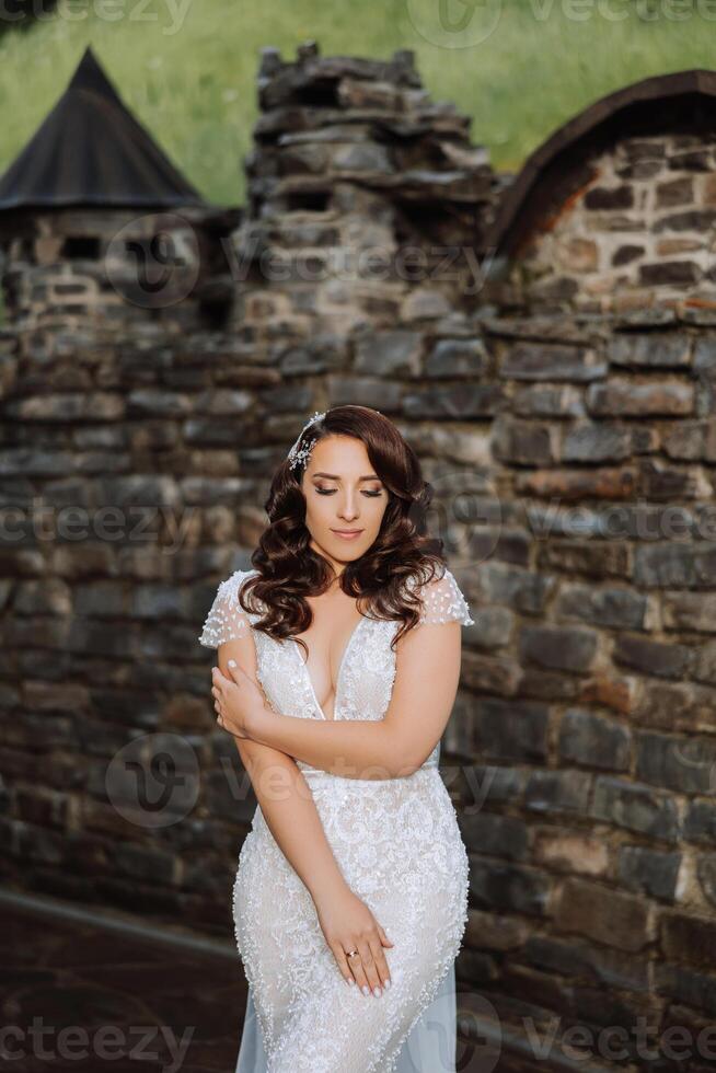 Curly brunette bride in a lace dress with an open bust, in a lush veil, poses against the background of green trees. Spring wedding photo