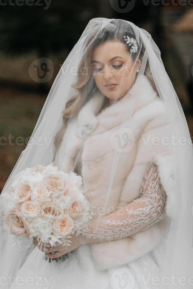 Portrait of a beautiful bride with a wedding bouquet of flowers, attractive woman in a wedding dress with a long veil. Happy bride woman. Bride with wedding makeup and hairdo. Winter wedding photo